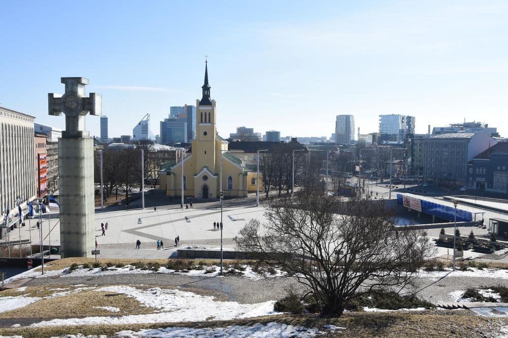 A cross-shaped stone monument overlooks a plaza with a yellow church spire and a modern skyline beyond